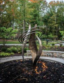 A modern sculpture with intertwined metal shapes stands surrounded by a fire pit, set in a landscaped garden with a backdrop of trees and foliage.