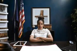 Woman sitting at a wooden desk with papers in front of her, crossing her arms with the american flag in the background.