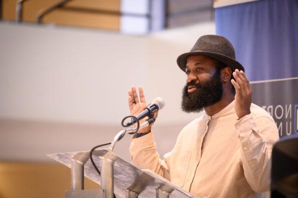 A man with a beard, wearing a hat and light-colored shirt, speaks into a microphone at a podium, urging everyone to stay connected.