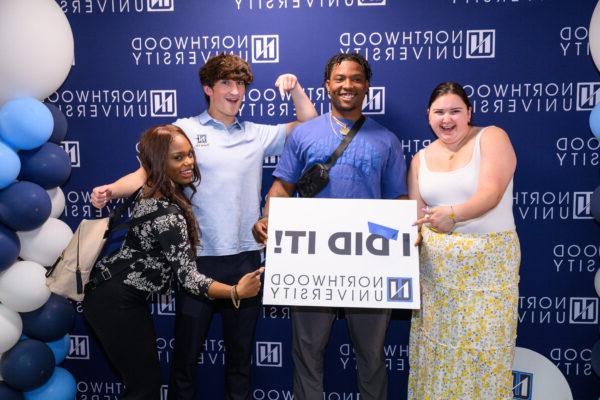 Four individuals pose in front of a 竞技宝app下载 University backdrop. One person holds a sign that reads "I DID IT!" Blue and white balloons are on the right side, celebrating their achievements and encouraging everyone to stay connected.