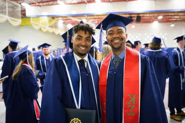 Two men in blue graduation gowns and caps, one wearing a red stole with Greek letters, smile at the camera at a graduation ceremony.