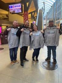 Four people holding Detroit Basketball sweatshirts pose inside a modern building with glass walls and stairs in the background, showcasing how fans can stay connected to their team.